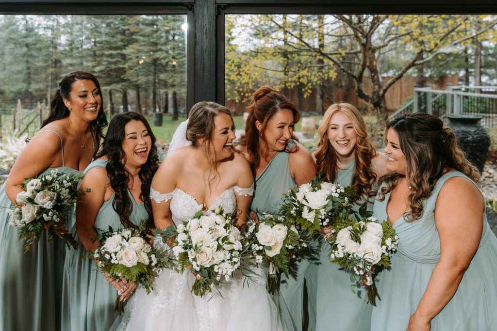 Bride laughing with bridesmaids in sage green dresses, all holding white floral bouquets