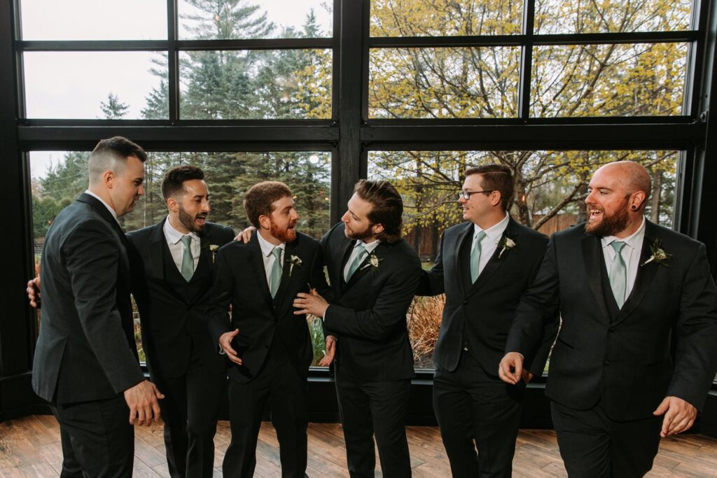 Groom and groomsmen joking around near large windows at Birch Wood Vineyards

