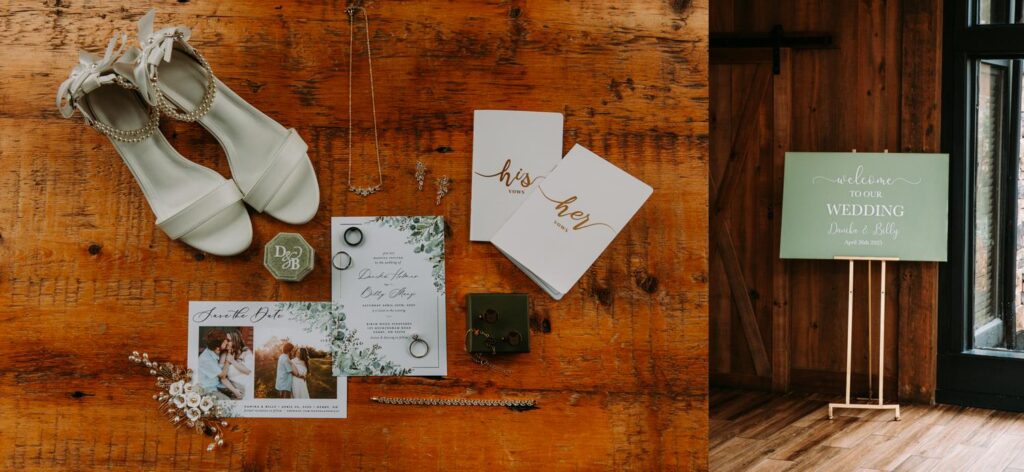 Flat lay of wedding details including shoes, invitations, vow books, and rings on rustic wooden table