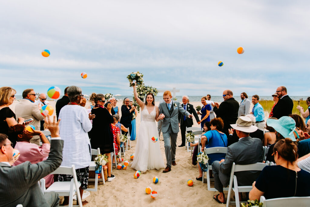 Newlyweds walking down the aisle at their Wychmere Beach Club wedding in Harwichport, Massachusetts, surrounded by guests tossing beach balls in celebration on the sand in a blog post for wedding day tips from a photographer