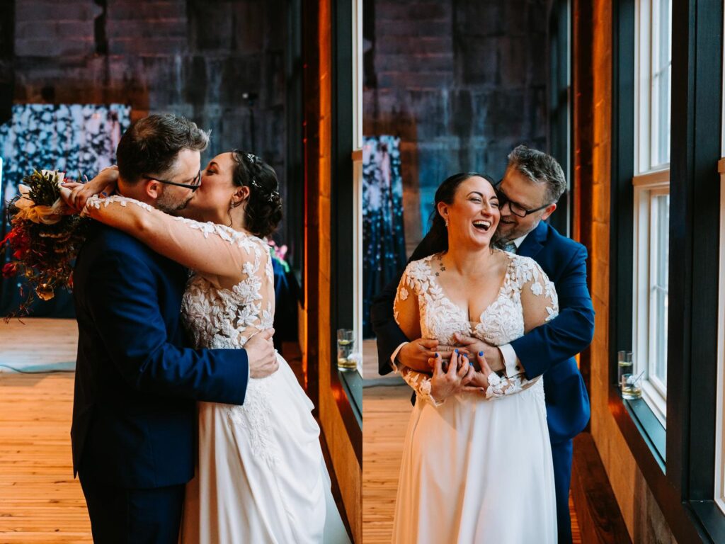 Groom wraps bride in an embrace, smiling in front of large factory-style windows.
