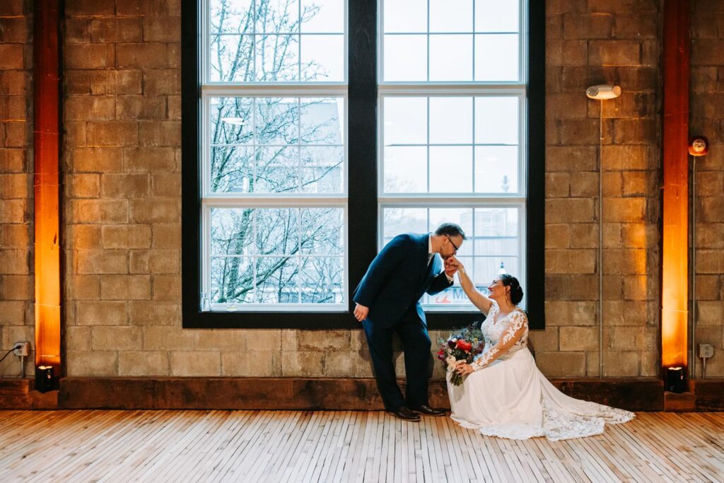 Groom kisses bride's hand in front of a warmly lit brick wall at Olio.