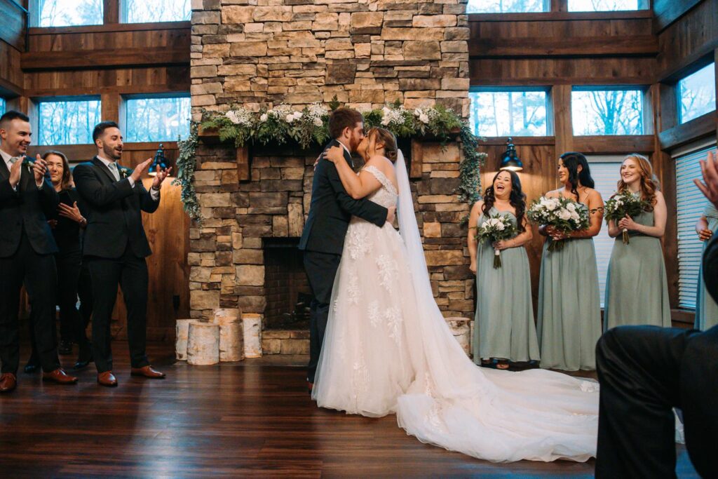 Full ceremony view as bride and groom share their first kiss with wedding party beaming in the background