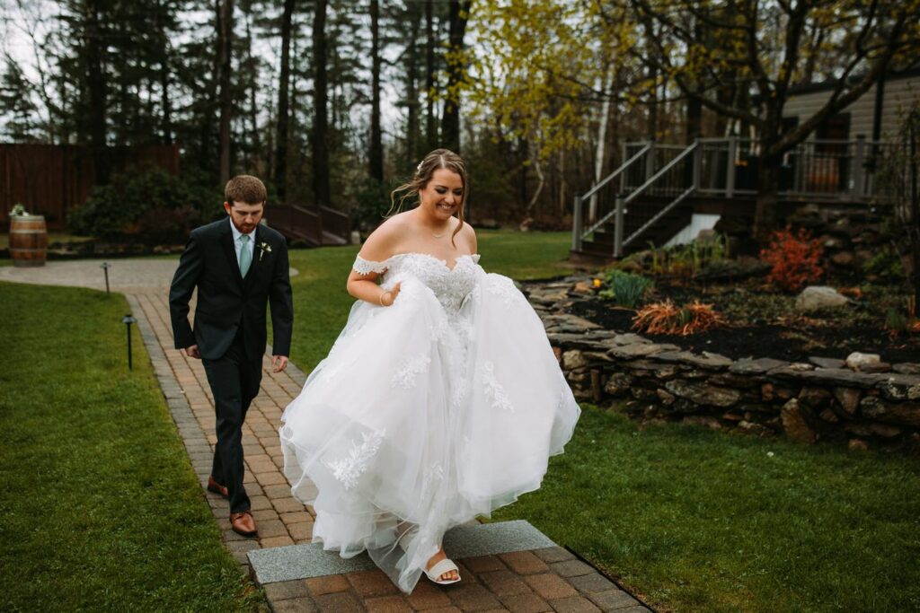 Bride smiling and lifting her dress slightly to walk across a brick path, with groom trailing behind her through the lush garden