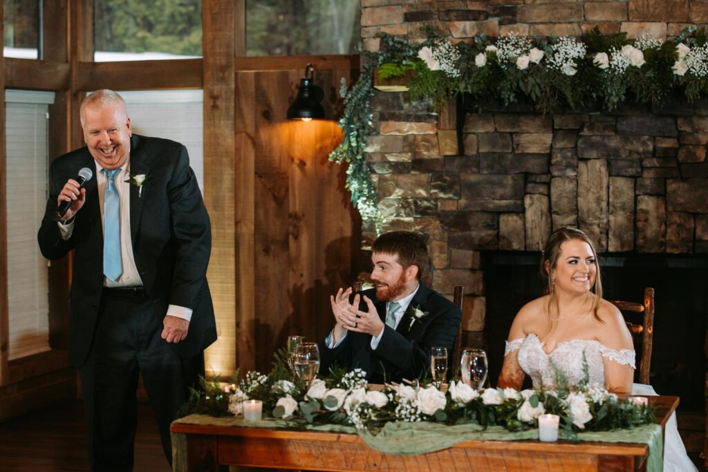 Father delivering a heartfelt toast while bride sits beside the stone fireplace backdrop