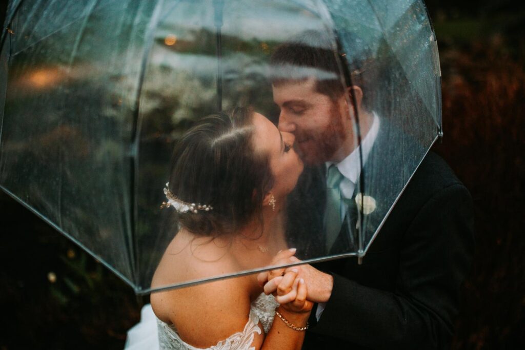 Bride and groom sharing a kiss under a clear umbrella, rain droplets visible on the surface

