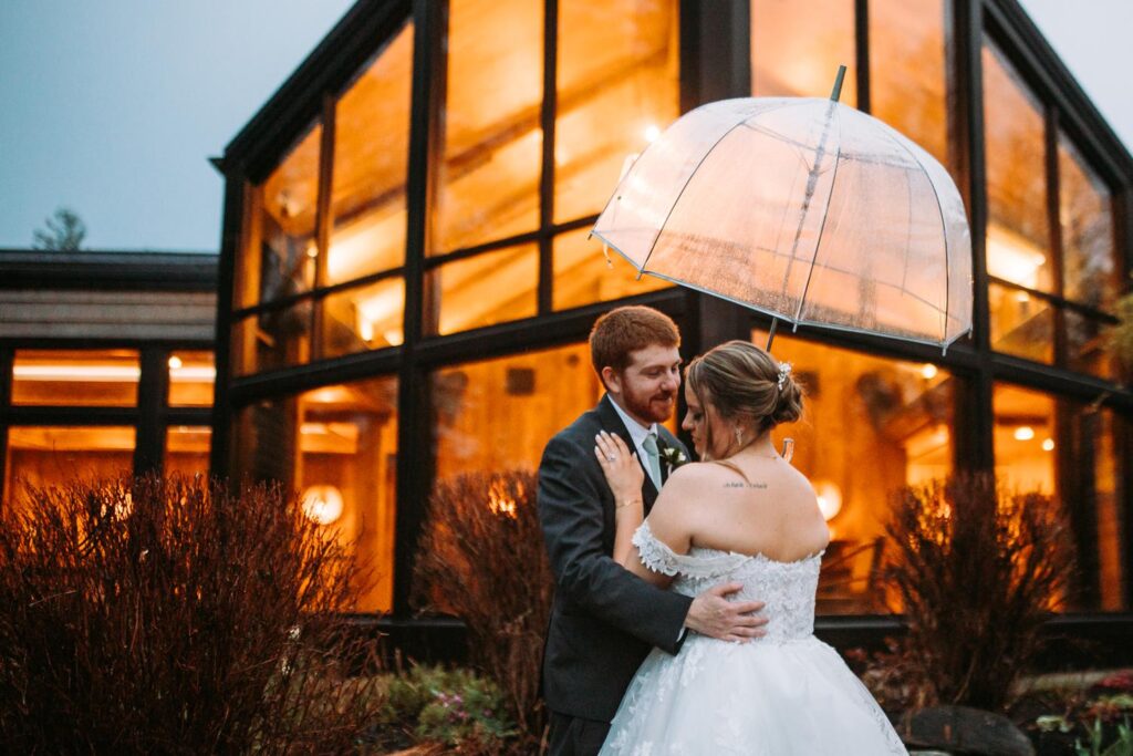 Groom embracing bride beneath a clear umbrella with the glowing venue behind them

