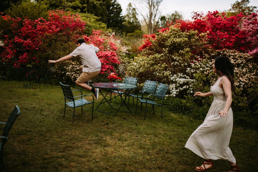 Groom-to-be goofily hopping toward a garden table while his fiancée laughs.