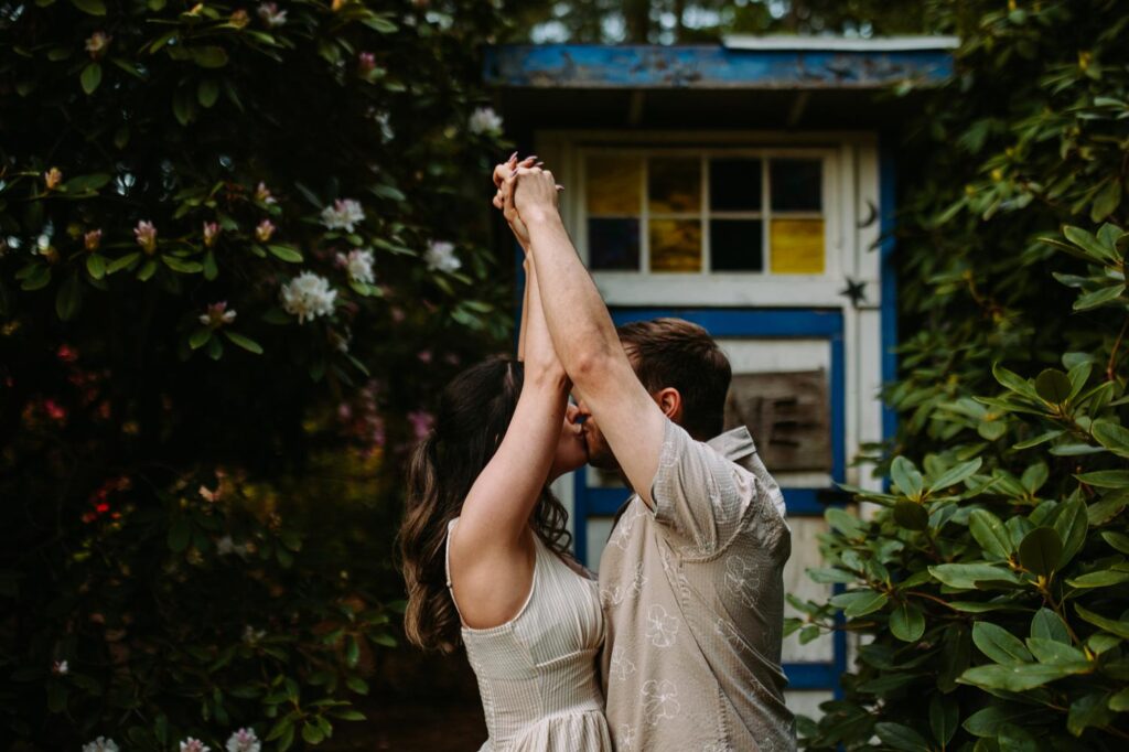 Couple kissing in front of a charming blue garden shed surrounded by greenery.