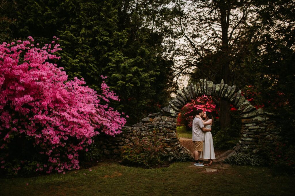 Couple embracing under Kinney Azalea Garden’s iconic stone arch, framed by glowing pink florals and towering trees at sunset.