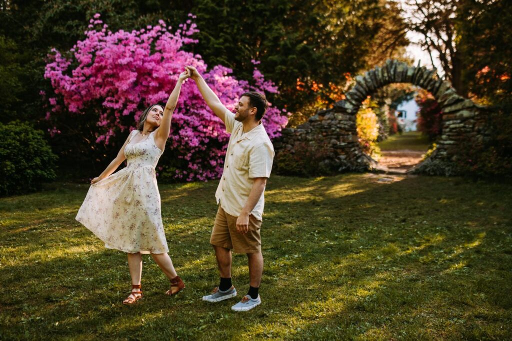 Couple twirling joyfully in front of bright pink azaleas and the stone arch in the distance.