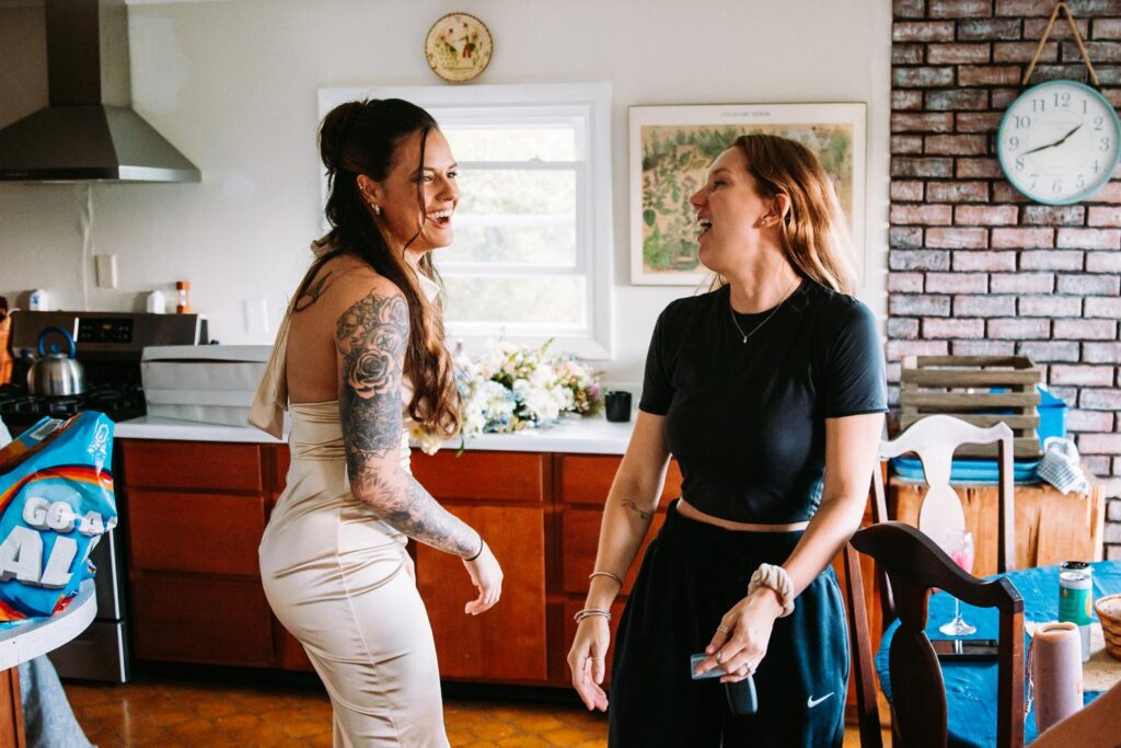 Bridesmaid and friend laugh together in the Bluebird Farm kitchen on wedding morning.