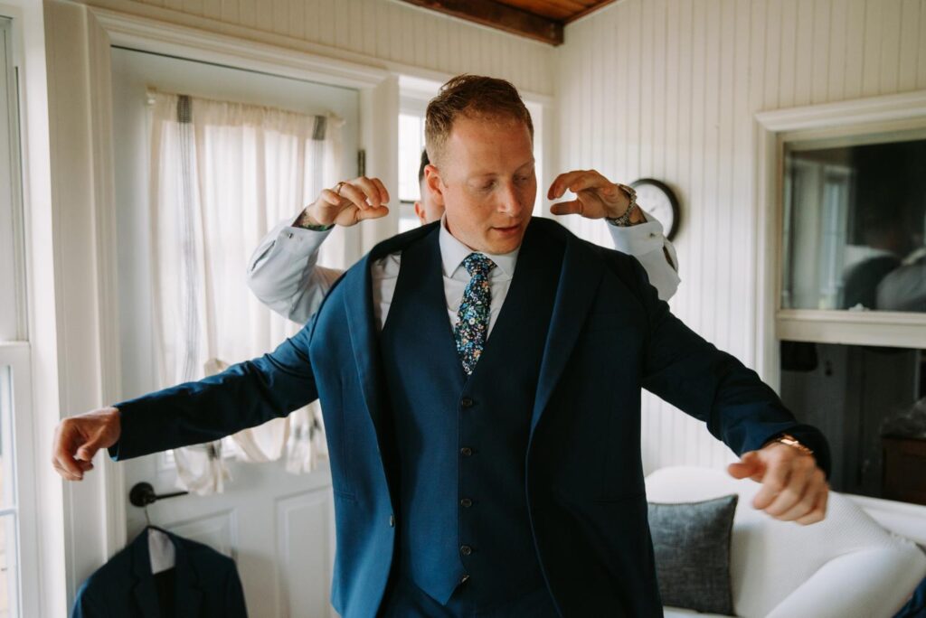 Groom adjusting suit jacket with help from groomsmen before Catskills wedding.