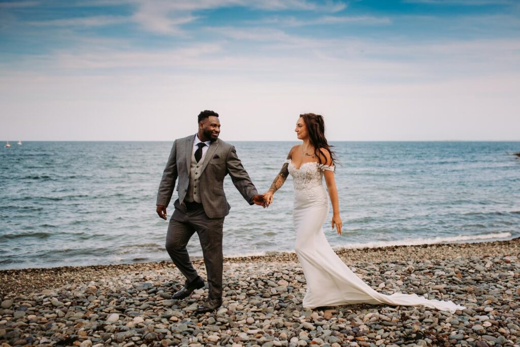 A bride and groom stroll along the rocky beach at their wedding at Oceanview of Nahant