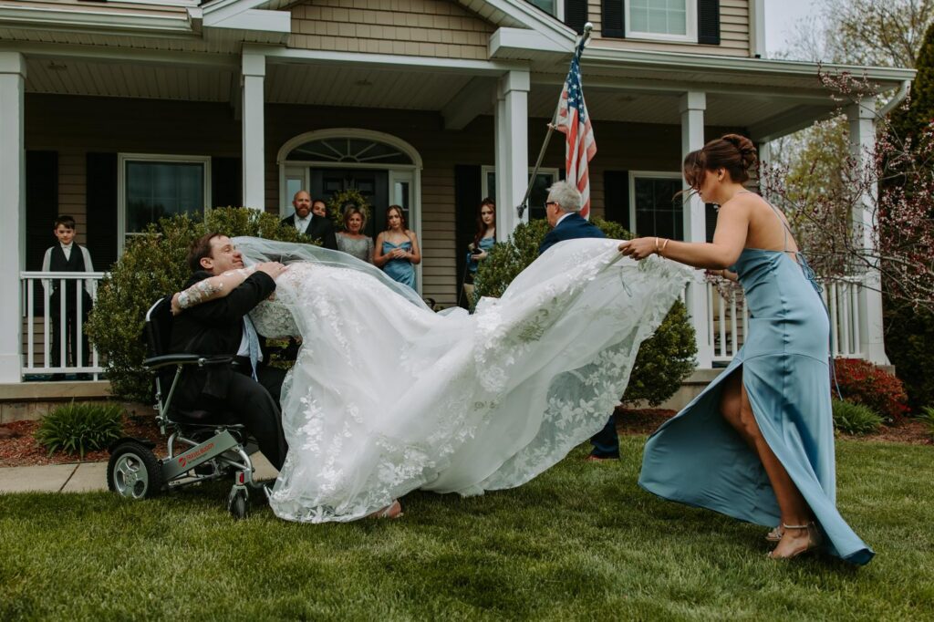 Bridesmaid fluffing and carrying bride's long lace train across the lawn before the ceremony