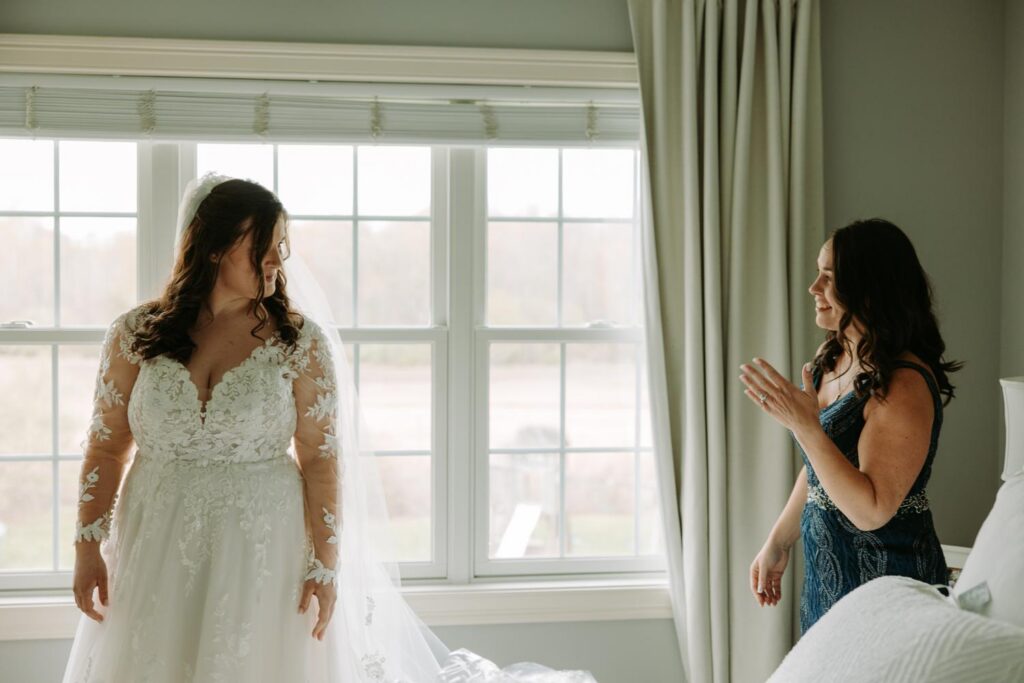 Bride in lace wedding dress standing at window with natural light during wedding morning preparations