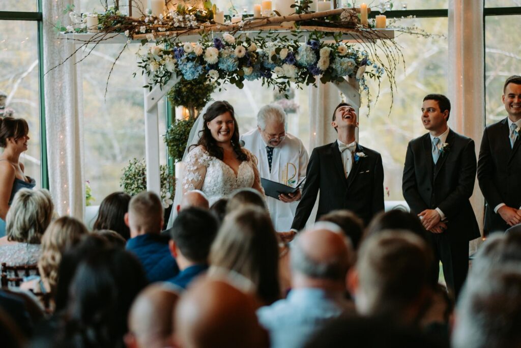 Wedding ceremony from behind showing guests, floral arch, and cathedral windows at The Pond House