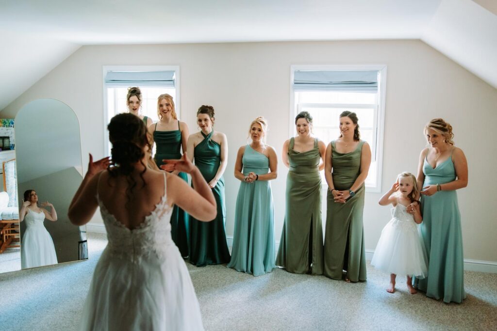 Bride from behind facing her bridesmaids in sage green dresses near a bright window before the ceremony