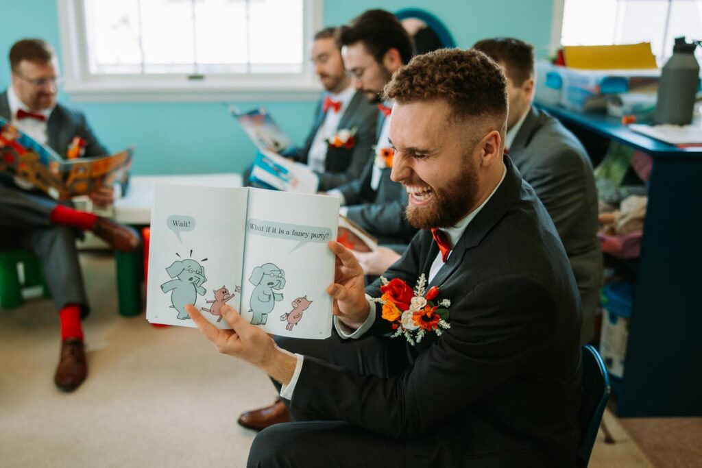 Groom laughing while opening a children's book gift surrounded by his groomsmen, red bow ties and suspenders, getting ready at Bluebird Farm