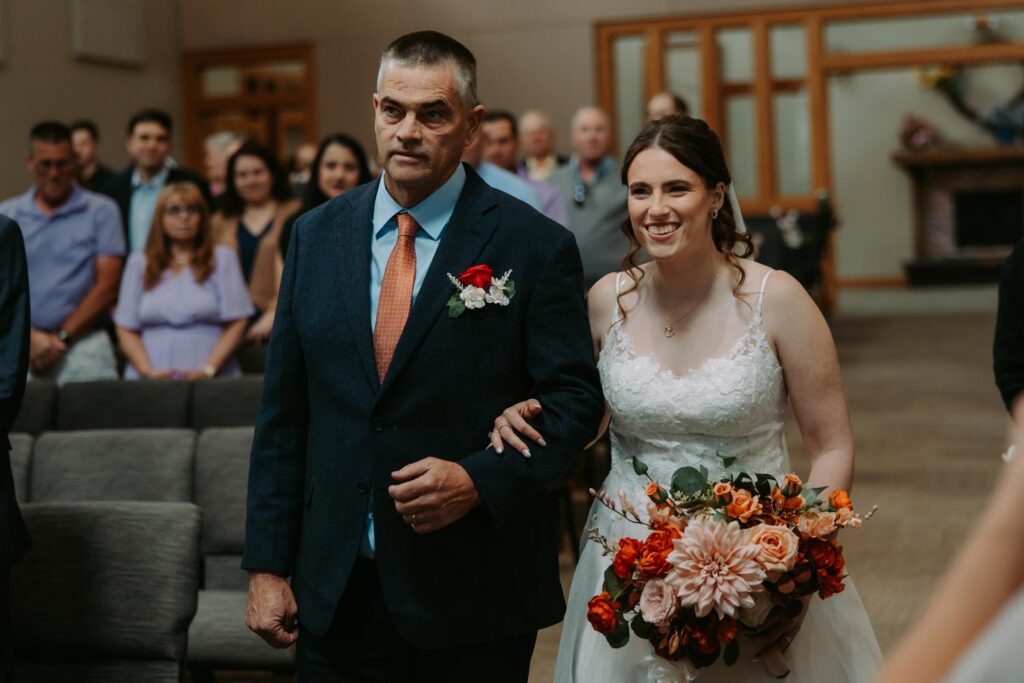 Father of the bride walking his daughter down the aisle, bride carrying orange and red dahlia bouquet, church ceremony