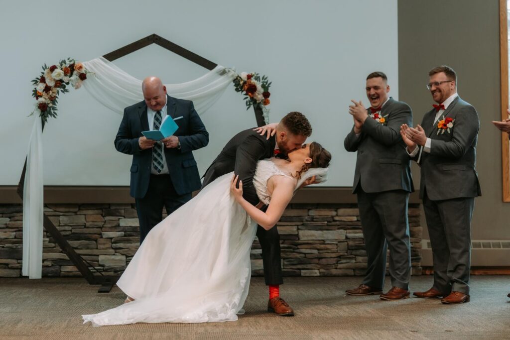 Bride and groom sharing their first kiss at the altar, guests applauding, wooden arch and sage green bridesmaids visible