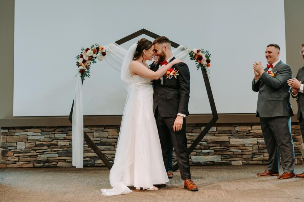 Bride and groom at the altar, full length shot, sage green bridesmaids and groomsmen in suspenders framing them