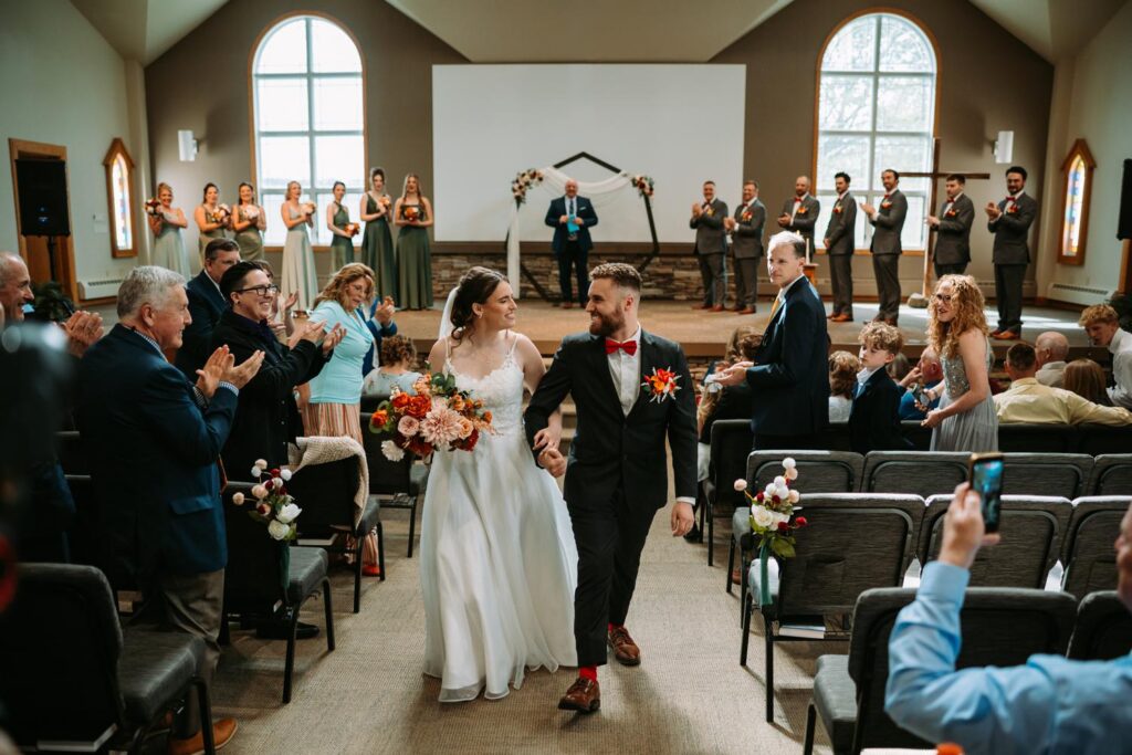 Wide shot of bride and groom walking back down the aisle after the ceremony, guests rising and cheering