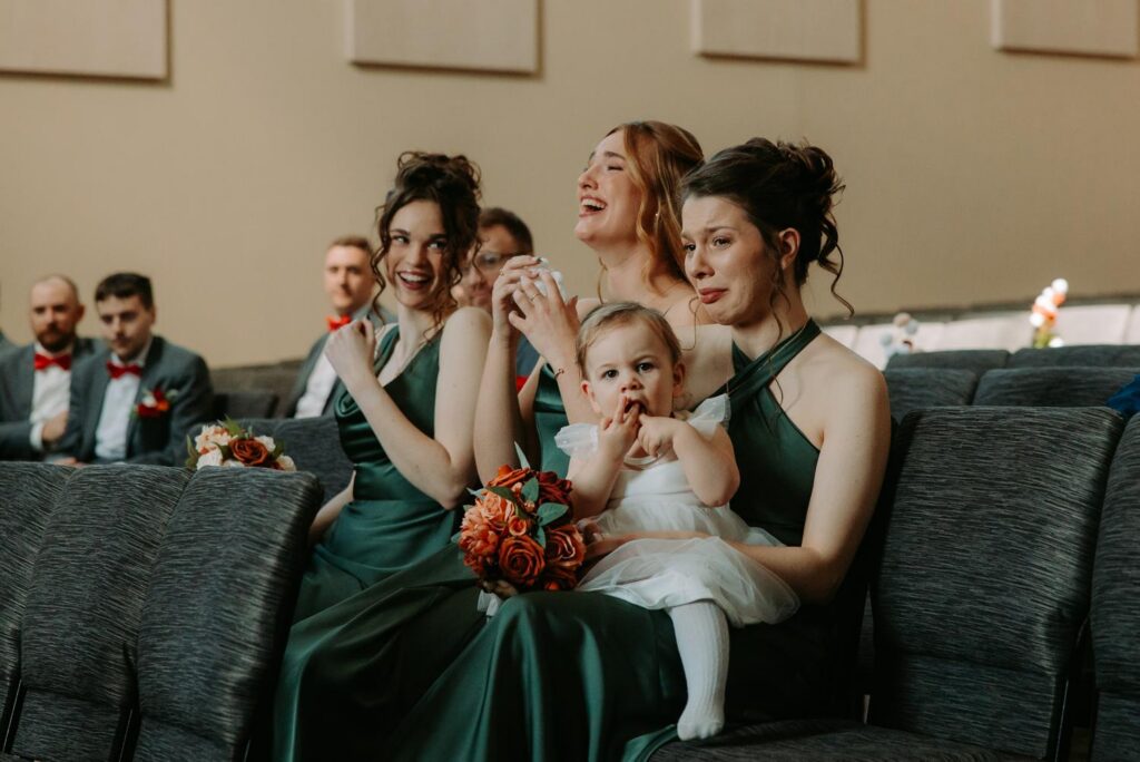 Bridesmaids in sage green dresses watching Tim's piano surprise with a baby in arms, emotional and joyful reactions