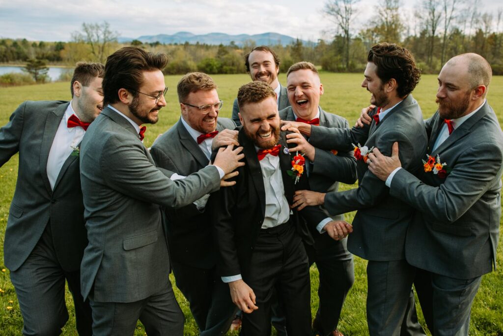 Groomsmen in suspenders and red bow ties laughing together in the field, groom in the center, Bluebird Farm wedding party