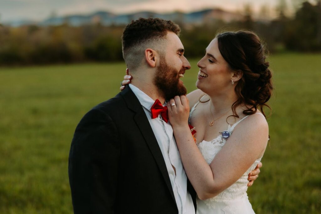 Close up of bride and groom laughing together in the field during golden hour portraits, red chili pepper boutonniere visible