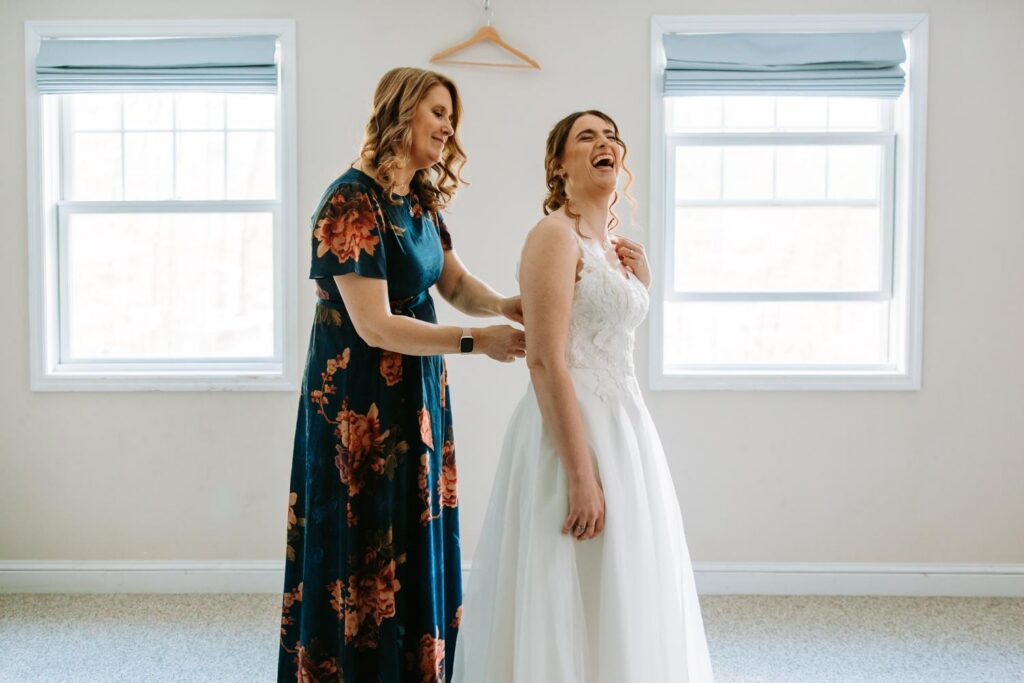 Bride in wedding dress laughing with a family member in a floral dress, getting ready room at Bluebird Farm wedding