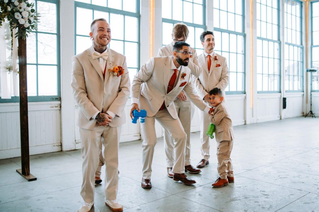 Groomsmen in tan suits laughing together inside Lighthouse Point Park venue, large windows with water view behind them