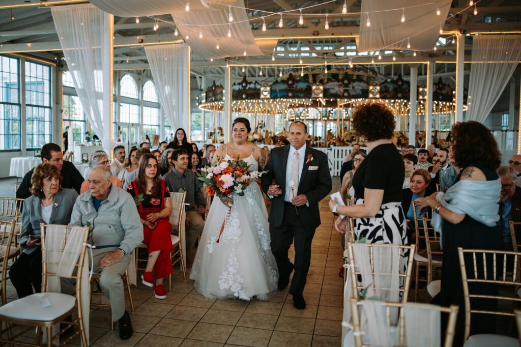 Wide shot of Nicole walking down the aisle at Lighthouse Point Park, full guest seating on both sides, string lights overhead