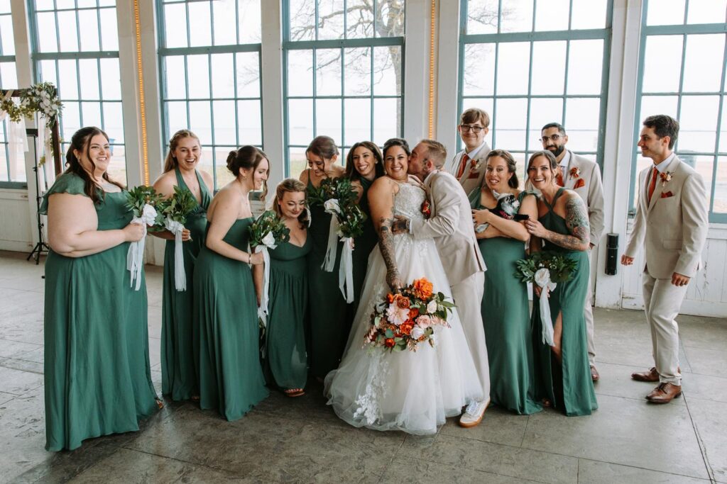 Full wedding party group laughing together in front of the large industrial windows at Lighthouse Point Park