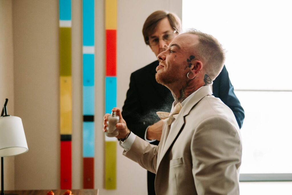 Sabin in tan suit getting sprayed with cologne in front of colorful striped wall, getting ready for his Lighthouse Point Park wedding