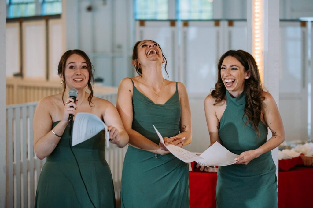 Bridesmaids in teal dresses giving a speech together at the microphone, both laughing, Lighthouse Point Park reception